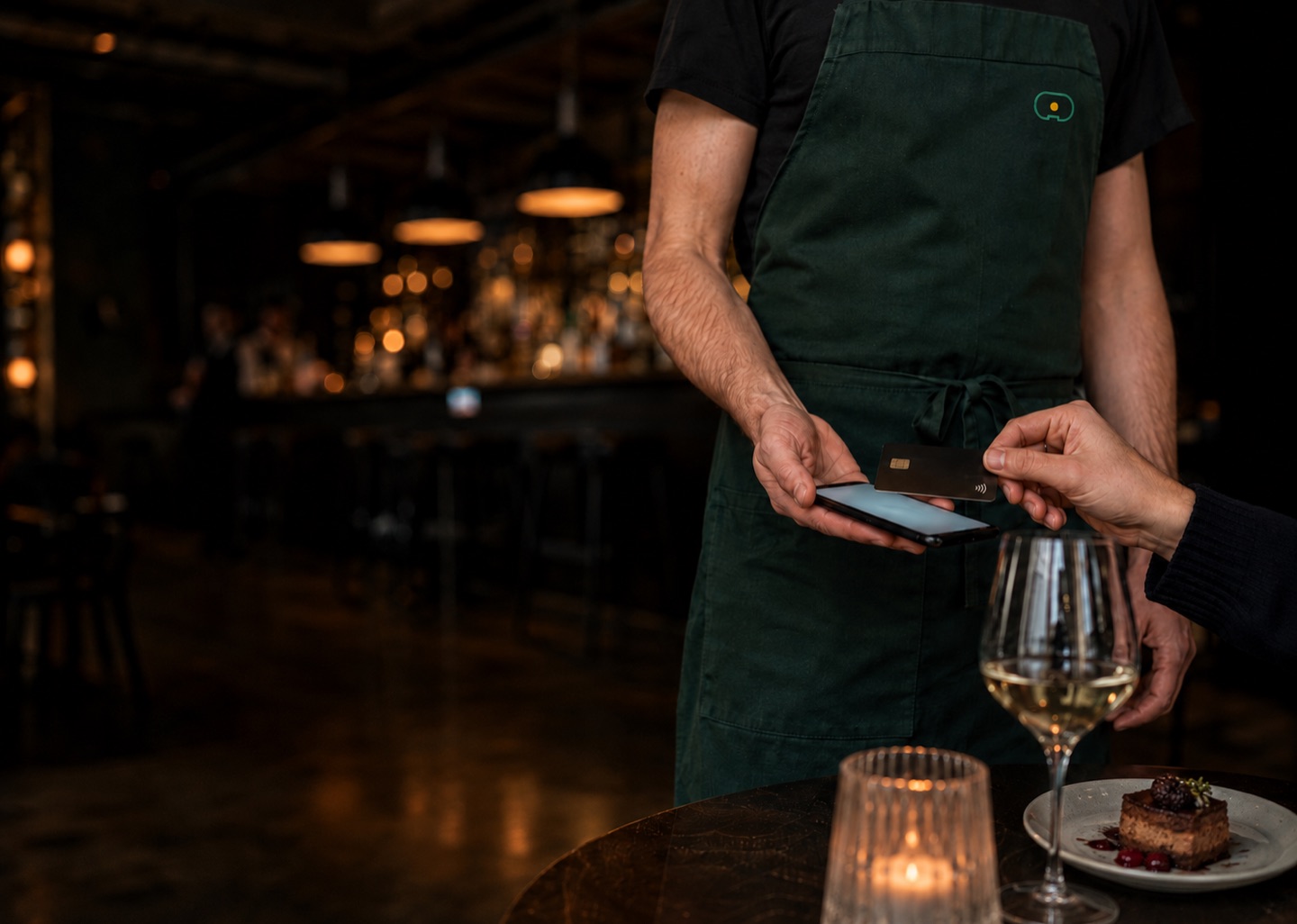 A server holding a phone at a table while a guest taps a card against it.